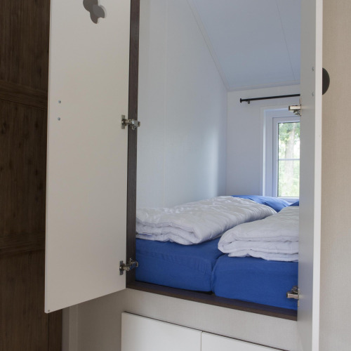 Helles Schlafzimmer in einem Naturhaus mit blauer Bettwäsche, weißen Decken und einem Fenster zur Natur.
