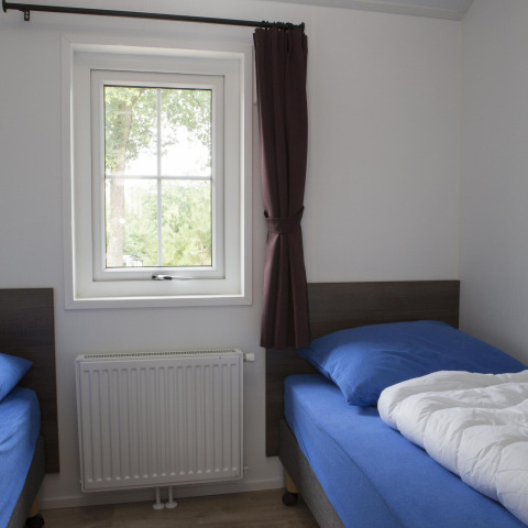 Habitación con dos camas individuales, ropa de cama azul, ventana con cortina oscura y radiador en casa natural.