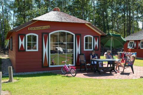 Casa de campo roja con terraza, tres mujeres, una niña, bicicleta rosa y sombrilla verde en el césped.