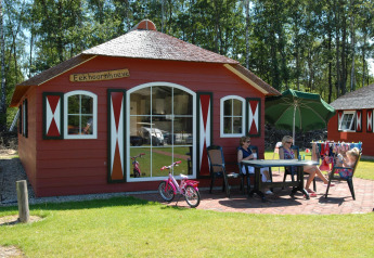Casa de campo roja con terraza, tres mujeres, una niña, bicicleta rosa y sombrilla verde en el césped.