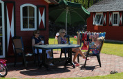 Two women and a child relax at a table outside a red nature house, with swimsuits drying on a clothesline.