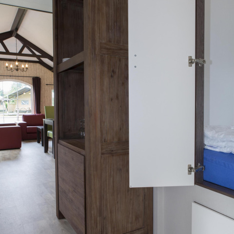 Interieur van een natuurhuisje met open kast, zitbanken en een groot raam met zicht op de tuin.