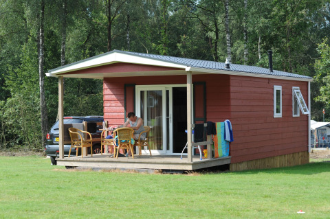 Cabaña roja de madera con porche y muebles de exterior en Veluwecamping De Pampel, rodeada de bosque.
