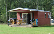 Cabaña roja de madera con porche y muebles de exterior en Veluwecamping De Pampel, rodeada de bosque.