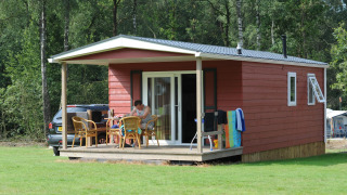Cabaña roja de madera con porche y muebles de exterior en Veluwecamping De Pampel, rodeada de bosque.