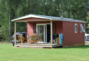 Cabaña roja de madera con porche y muebles de exterior en Veluwecamping De Pampel, rodeada de bosque.