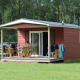 Rote Holzhütte mit Veranda und Gartenmöbeln auf der Veluwecamping De Pampel, umgeben von Wald.