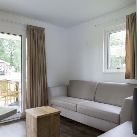 Stue i en Forest lodge på Veluwecamping De Pampel i Holland med sofa, træbord og udsigt til terrassen.
