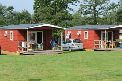 Zwei rote Hütten im Wald, Forest Lodge, Veluwecamping De Pampel in den Niederlanden, Menschen sitzen draußen.