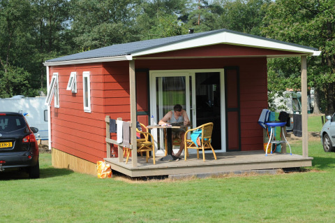 Cabaña roja con porche y hombre en la mesa, Forest lodge en Veluwecamping De Pampel, Países Bajos.