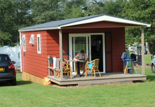 Rød lodge med veranda og mand ved bordet, Forest lodge ved Veluwecamping De Pampel i Holland.