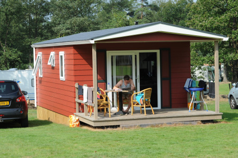 Iemand zit aan een tafel op de veranda van een rood huisje bij Forest lodge op Veluwecamping De Pampel.