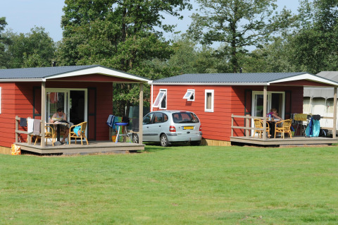Twee rode blokhutten met veranda, auto en mensen bij Veluwecamping De Pampel in Nederland.