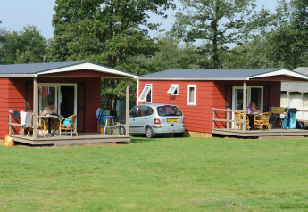 To røde skovhytter, bil og mennesker på veranda, græsplæne ved Veluwecamping De Pampel, Holland.