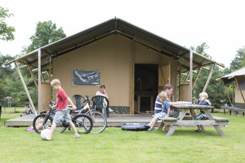 Famiglia si rilassa davanti a una tenda safari Woodlodge. Bambini giocano con biciclette, altri al tavolo da picnic.
