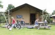 Family relaxing outside a Woodlodge safari tent. Children play with bikes while others sit at a picnic table.