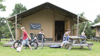 Famiglia si rilassa davanti a una tenda safari Woodlodge. Bambini giocano con biciclette, altri al tavolo da picnic.