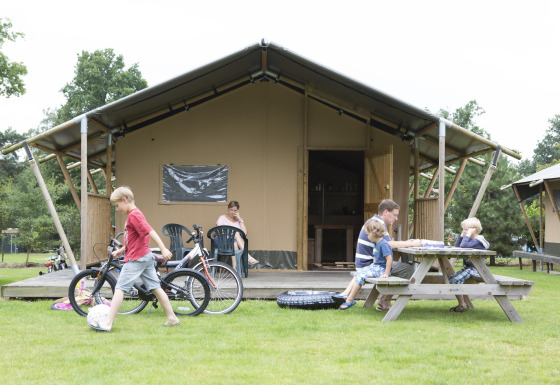 Family relaxing outside a Woodlodge safari tent. Children play with bikes while others sit at a picnic table.