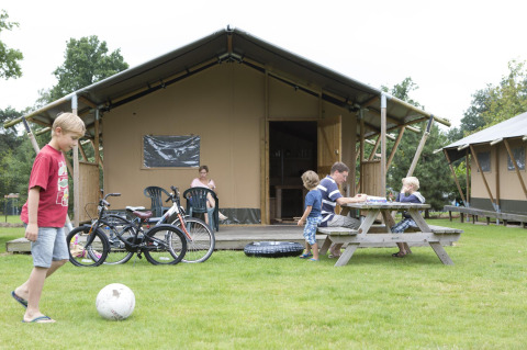 Famille devant une tente safari Woodlodge, enfants jouent, vélos garés et pique-nique sur l’herbe.