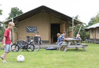 Familia disfrutando frente a una tienda safari Woodlodge, niños juegan y bicicletas en césped verde.