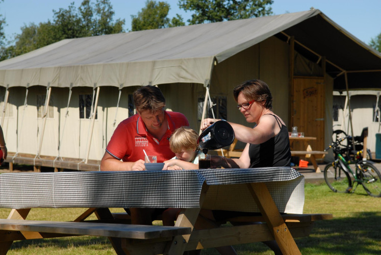 Family having an outdoor meal at a picnic table in front of a safari tent called Woodlodge in sunlight.