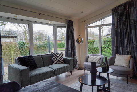 Moderne woonkamer in een vakantiehuis met grote ramen, grijze zetel en stoelen bij EuroParcs Noordwijkse Duinen.