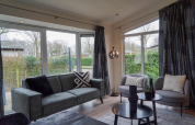 Moderne woonkamer in een vakantiehuis met grote ramen, grijze zetel en stoelen bij EuroParcs Noordwijkse Duinen.