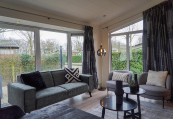 Moderne woonkamer in een vakantiehuis met grote ramen, grijze zetel en stoelen bij EuroParcs Noordwijkse Duinen.