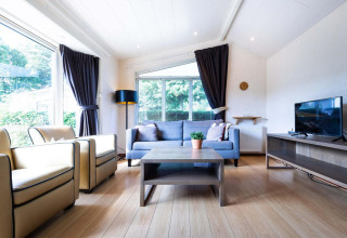 Lyst opholdsområde i træhytte på EuroParcs Noordwijkse Duinen med store vinduer, sofa og tv.