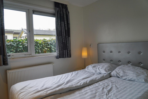 Slaapkamer met tweepersoonsbed, lampje en raam met zicht op tuin bij EuroParcs Noordwijkse Duinen.