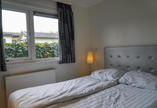 Slaapkamer met tweepersoonsbed, lampje en raam met zicht op tuin bij EuroParcs Noordwijkse Duinen.