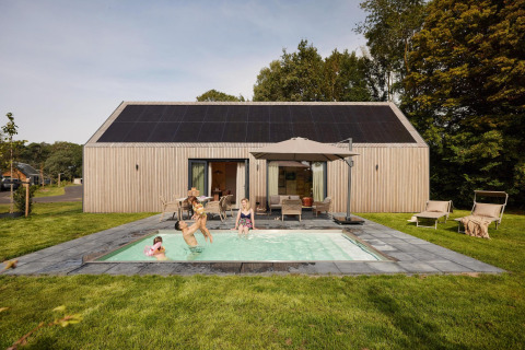 Una familia disfruta en una piscina exterior frente a una moderna cabaña con paneles solares en el tejado.