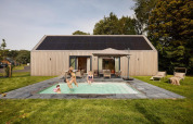 Una famiglia si diverte in una piscina esterna davanti a un lodge moderno con pannelli solari sul tetto.