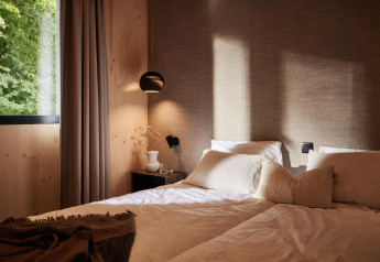 Knusse lodge slaapkamer met zachte kussens, houten muren en groot raam met uitzicht op groen bos.