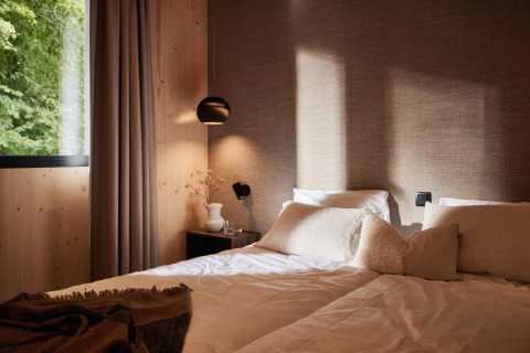 Knusse lodge slaapkamer met zachte kussens, houten muren en groot raam met uitzicht op groen bos.