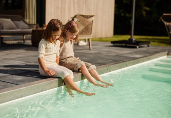 Due bambine sono sedute sul bordo di una piscina in un lodge e immergono i piedi nell’acqua, al sole.