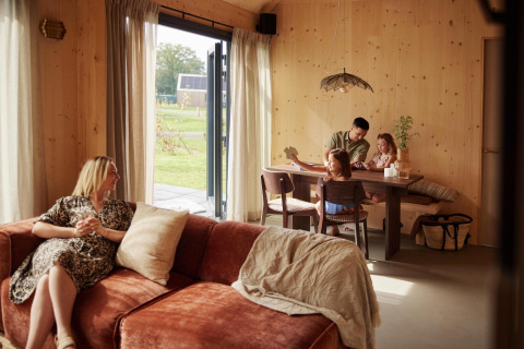 A family enjoys time inside a modern lodge with wooden walls; a woman relaxes on the couch while others play cards.