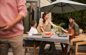 Familie sitzt gemeinsam beim Essen im Freien unter einem Sonnenschirm an einer Lodge, lacht fröhlich.