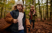 Un enfant court dans une forêt d’automne près d’un lodge, un adulte portant un enfant en arrière-plan.