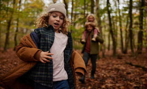 Een kind loopt in een herfstbos bij een lodge, terwijl een volwassene achteraan een kind op de schouders draagt.