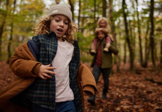 Un niño corre por un bosque otoñal cerca de una cabaña, mientras un adulto lleva a otro niño a cuestas atrás.