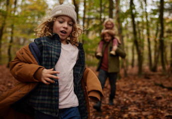 Un niño corre por un bosque otoñal cerca de una cabaña, mientras un adulto lleva a otro niño a cuestas atrás.