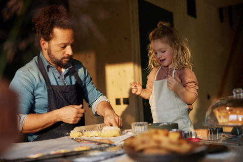 Padre e hija hornean juntos en la cocina cálida de un lodge, ambos con delantales y sonriendo felices.