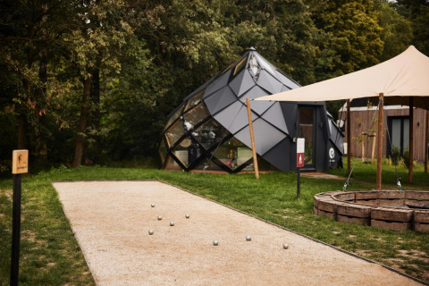 Lodge moderno de glamping con diseño geométrico, pista de petanca y fogata en entorno natural verde.