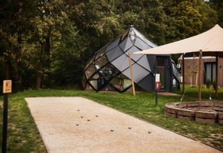 Lodge moderno de glamping con diseño geométrico, pista de petanca y fogata en entorno natural verde.
