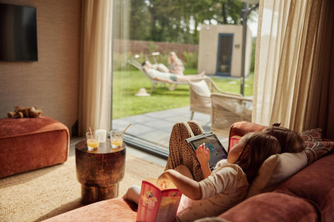 Kind ligt op de zetel met tablet terwijl volwassenen buiten genieten in de tuin bij Unbrick One Family-lodge.