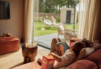 Kind ontspant op de bank met tablet, terwijl volwassenen genieten in de tuin bij Unbrick One Family-lodge.