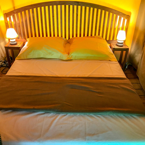 Cama cómoda con cabecero de madera, lámparas y mesas laterales dentro de la tienda safari Lodgetent Gourdon en Francia.