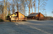Dos cabañas de madera en River Lodge, Camping de Koeksebelt, Países Bajos, rodeadas de césped y árboles altos.