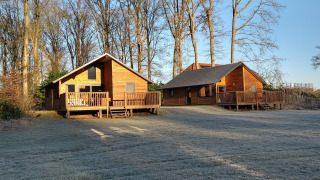Two wooden cabins at River Lodge, Camping de Koeksebelt, Netherlands, with frost on the grass and tall trees.